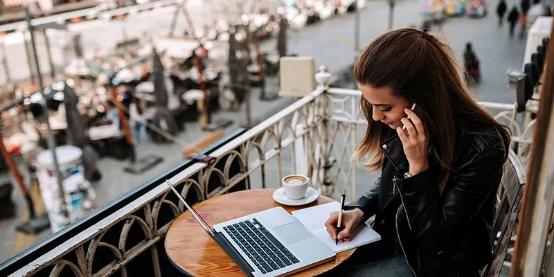 Mujer trabajando con portátil desde terraza urbana mientras habla por teléfono