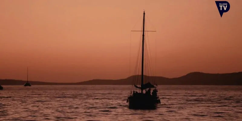 Velero navegando al atardecer con cielo anaranjado y siluetas en el horizonte