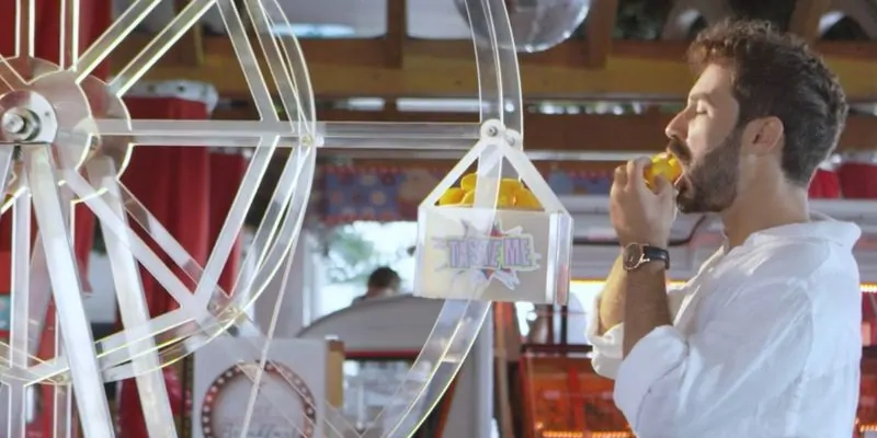 Hombre degustando fruta tropical frente a rueda decorativa en espacio estilo feria