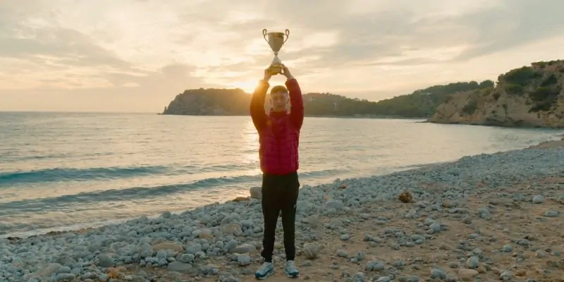 Niño celebrando con trofeo al atardecer en una playa rocosa