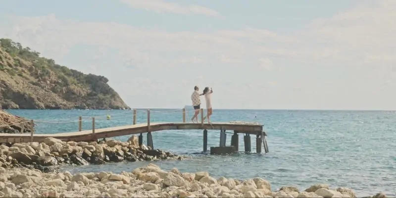 Pareja caminando por un muelle de madera sobre aguas turquesas en costa rocosa